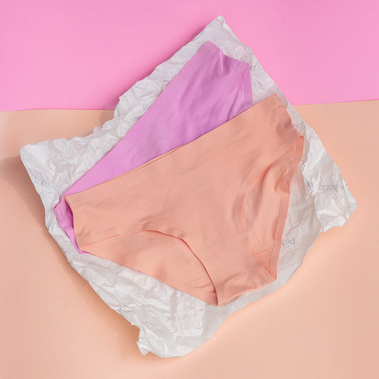 Two pairs of underwear, one pink and one peach, on a white lace background with a pink and beige gradient.