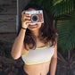 Person holding a vintage camera in front of their face with plants in the background wearing Qiwion jumpstart bra in a light grey, with neon straps and underband