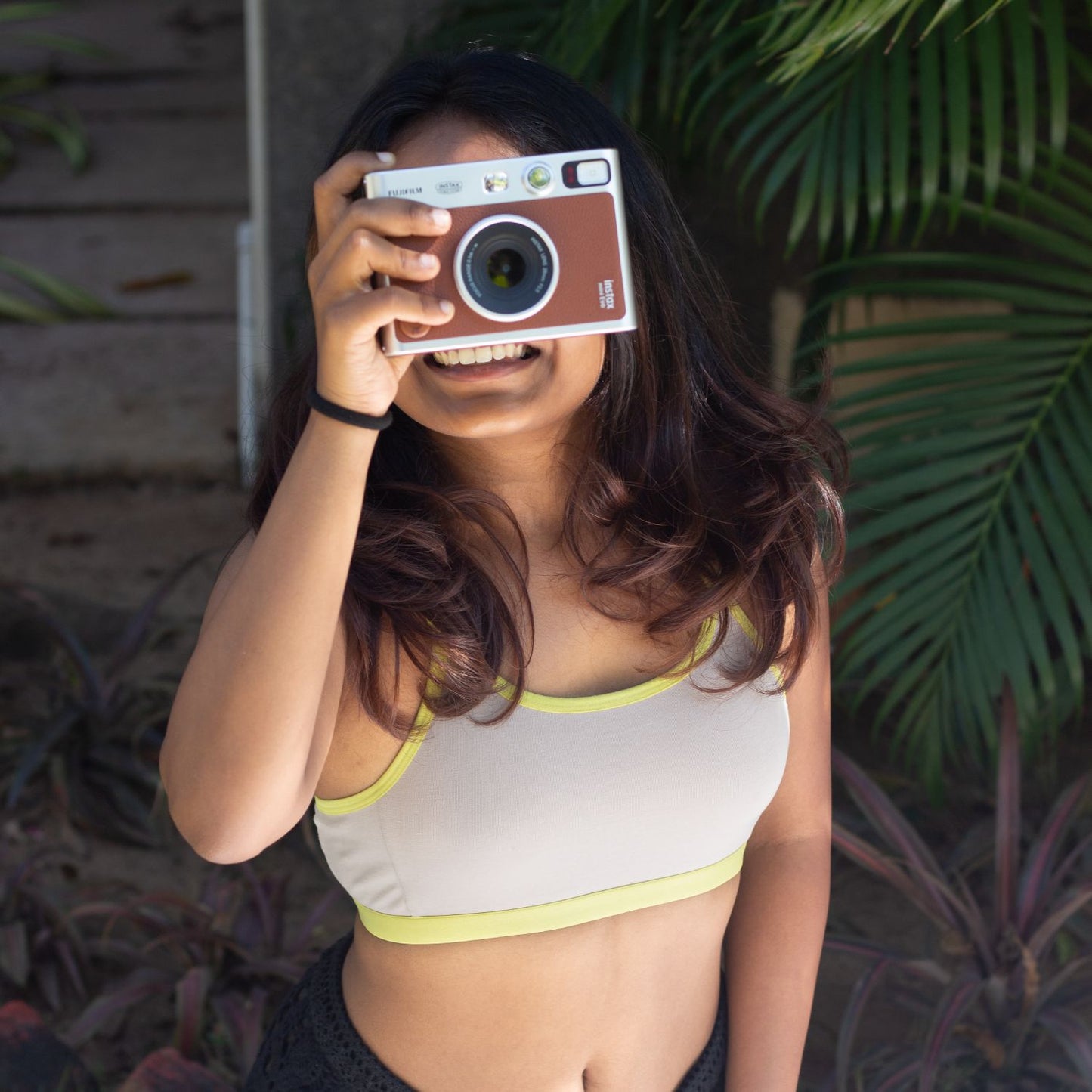 Person holding a vintage camera in front of their face with plants in the background wearing Qiwion jumpstart bra in a light grey, with neon straps and underband