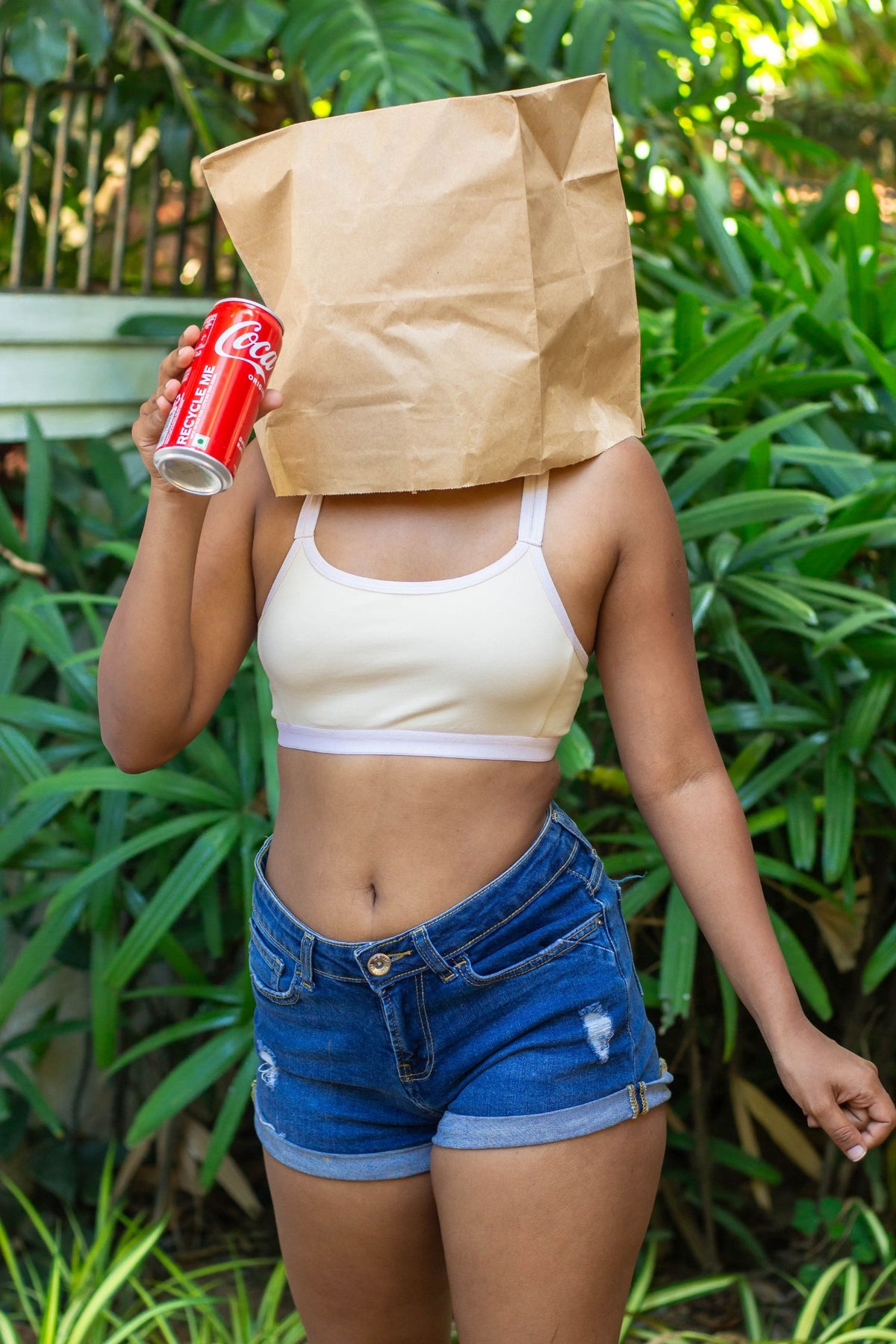 Person holding a Coca-Cola can with a paper bag over their head outdoors, wearing a qiwion jumpstart bra in cream colour, with a pair of shorts. 
