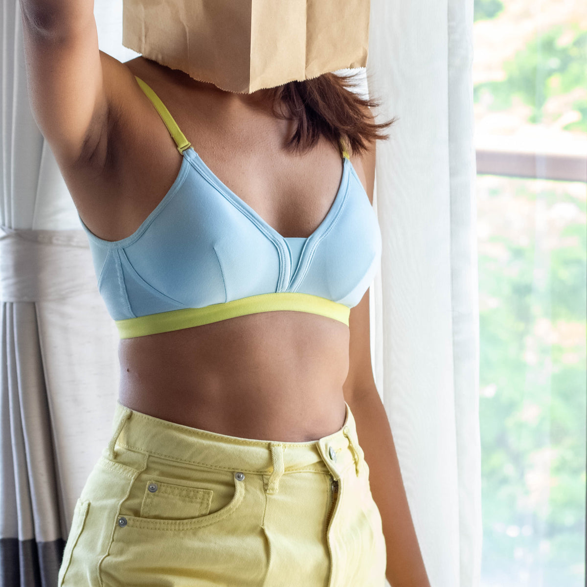 Person wearing a light blue qiwon bra and yellow pants with a paper bag over their head indoors.