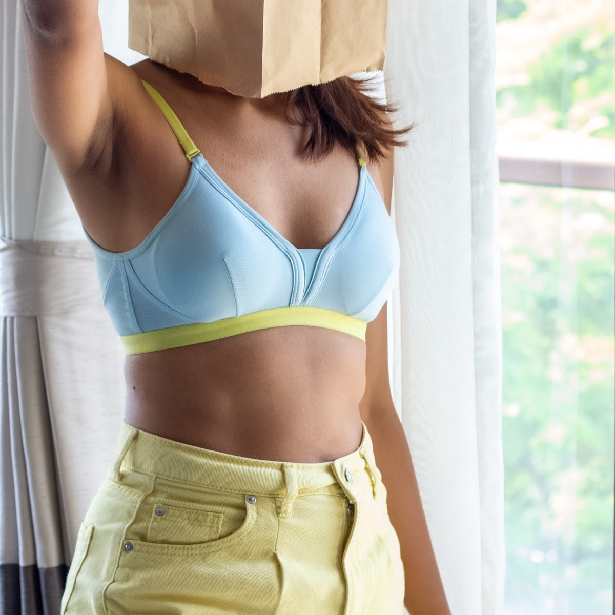 Person wearing a light blue bra and yellow pants with a paper bag over their head indoors.