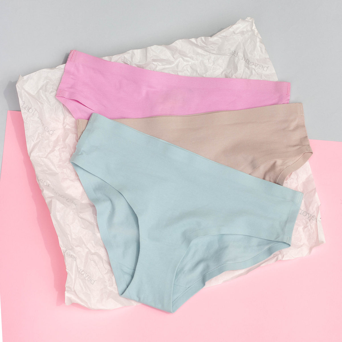 Three pairs of underwear in pink, blue, and light grey on a pink surface with a gray background.