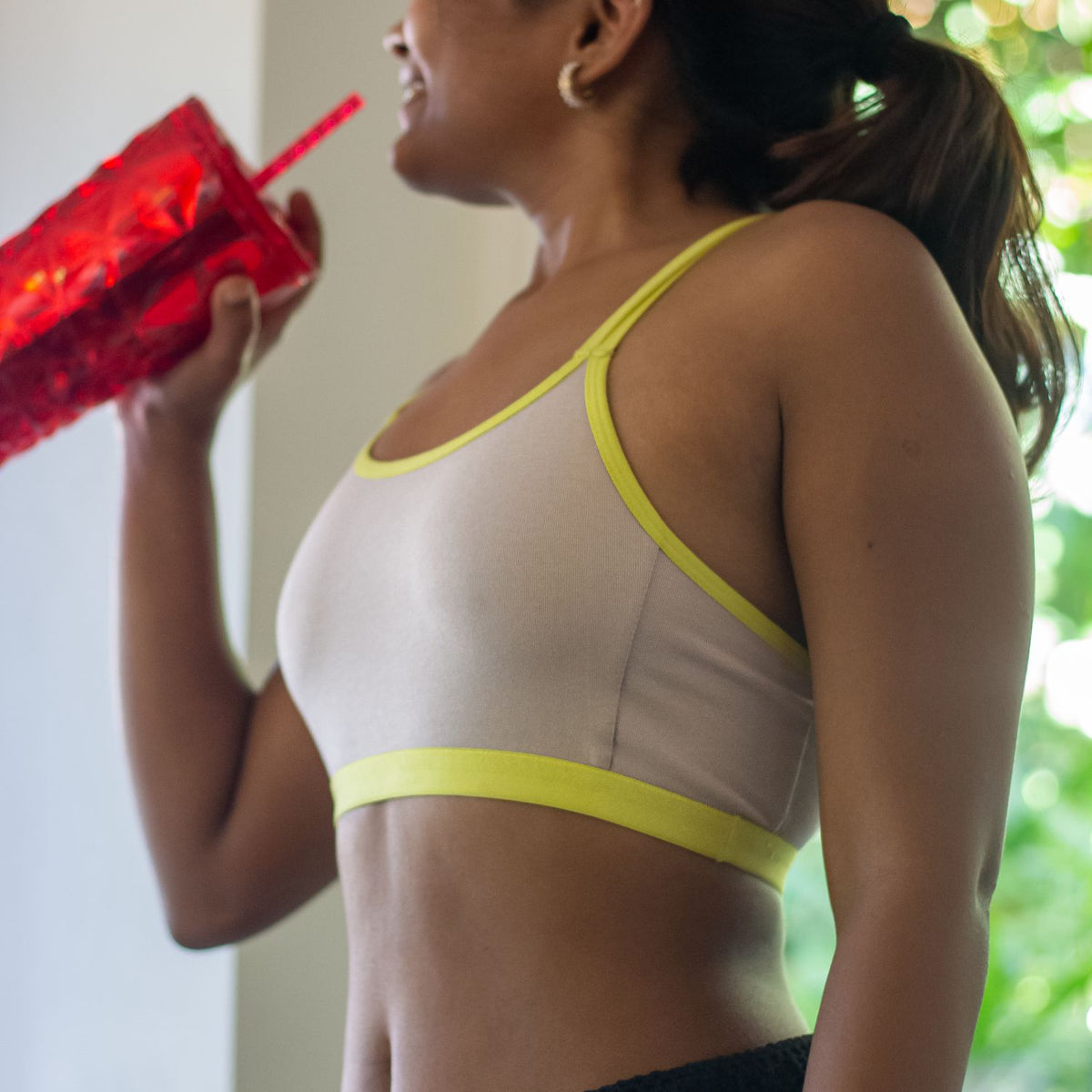 Woman wearing a qiwion jumpstart bra  in light grey and with neon straps holding a red drink container.