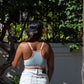 Person wearing a light blue sports bra and white pants standing in front of greenery.
