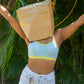 Person wearing a light blue qiwion bra and white pants with a brown paper bag over their head, standing against a green leafy background.