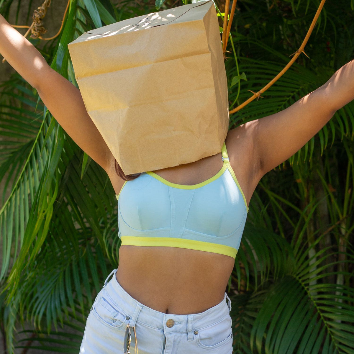 Person wearing a light blue qiwion bra and white pants with a brown paper bag over their head, standing against a green leafy background.