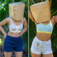 Two images of a person wearing a brown paper bag over their head with a smiley face, one in a white bra and blue shorts, the other in a light blue sports bra and white shorts, against a green leafy background.